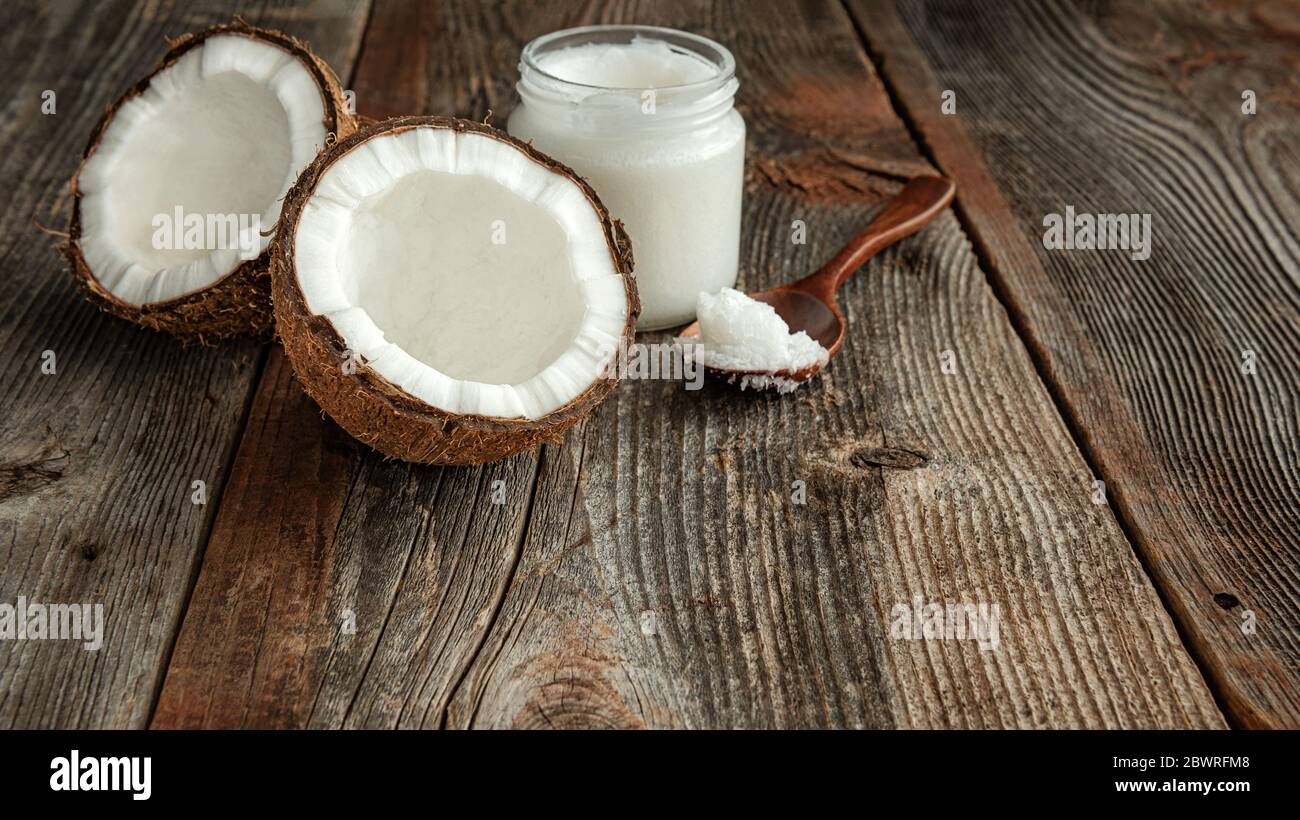 Due metà di cocco, cucchiaio e olio di cocco in un vaso su una superficie di legno. Il concetto di cibo sano. Foto Stock