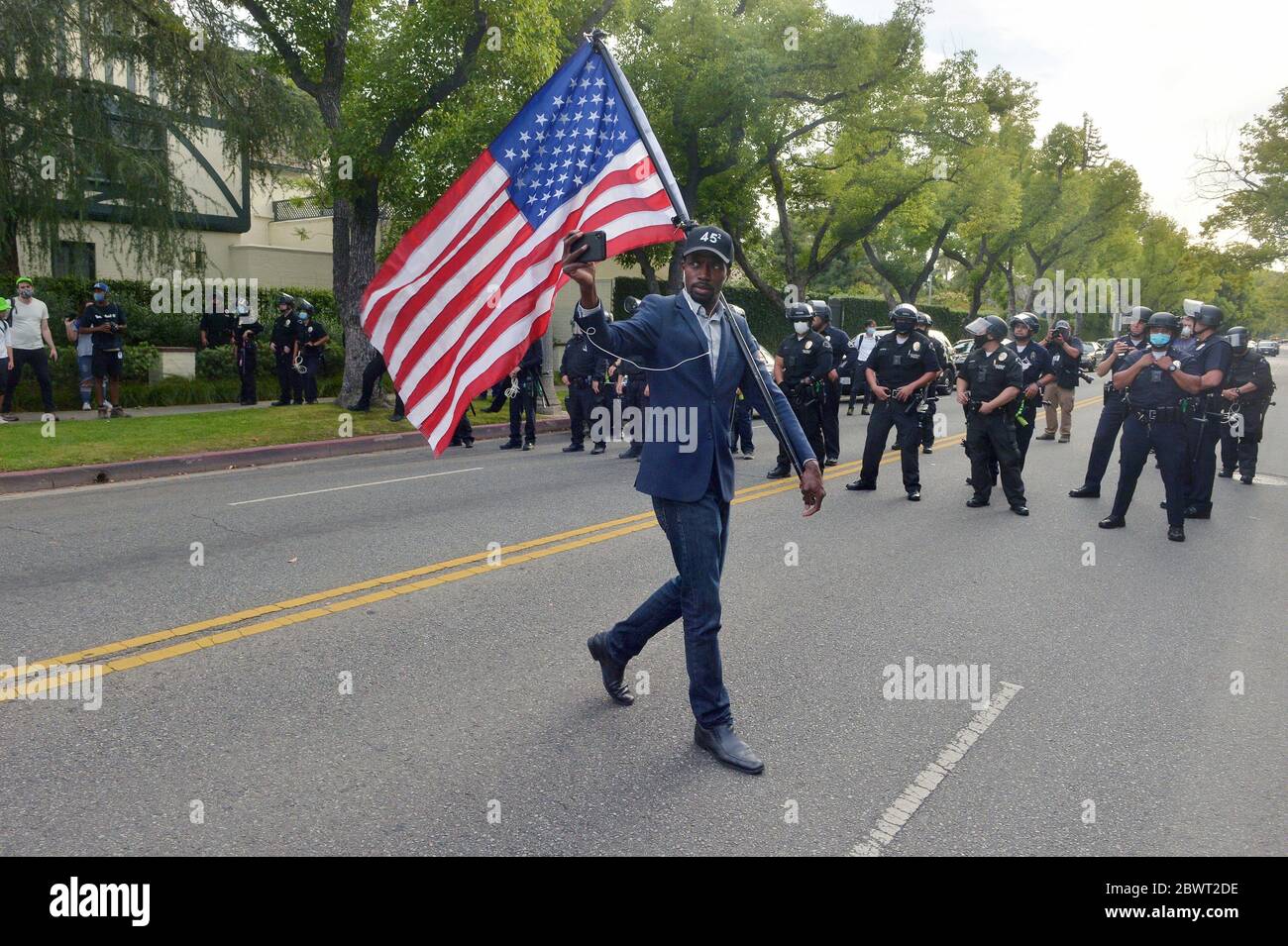 Un uomo passa davanti a ufficiali di polizia con una bandiera americana durante una protesta pacifica dell'uccisione di George Floyd a Minneapolis nella residenza ufficiale del sindaco Eric Garcetti nella sezione Hancock di Los Angeles martedì 2 giugno 2020. Il presidente Trump ha minacciato lunedì sera di inviare l'esercito degli Stati Uniti in qualsiasi città in cui i funzionari locali non riescono a controllare folle di manifestanti indisciplinati. Il suo segretario di difesa, Mark T. Esper, si è scagliato sulla necessità di truppe per 'dominare lo spazio di battaglia' nelle città americane. Trump sembra aver intenzione di invocare l’Insurrection Act. Foto di Jim Ruy Foto Stock