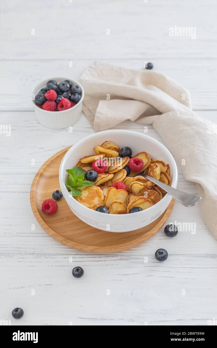 Prima colazione ciotola bianca con minuscoli cereali pancake con mirtilli, lamponi e menta su sfondo di legno Foto Stock