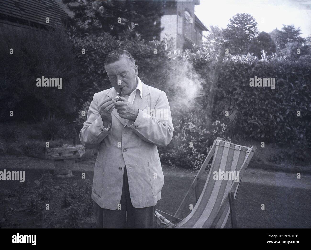 Anni '50, storico, un Signore inglese che indossa una giacca di tela in piedi presso la sua sedia a sdraio all'esterno in un giardino che illumina il suo pipe, Inghilterra, Regno Unito. Il fumo di pipa è la forma più antica e più tradizionale di fumo, più comunemente tabacco ed era particolarmente popolare tra i gentiluomini di una certa età fino ai primi anni 60. Foto Stock