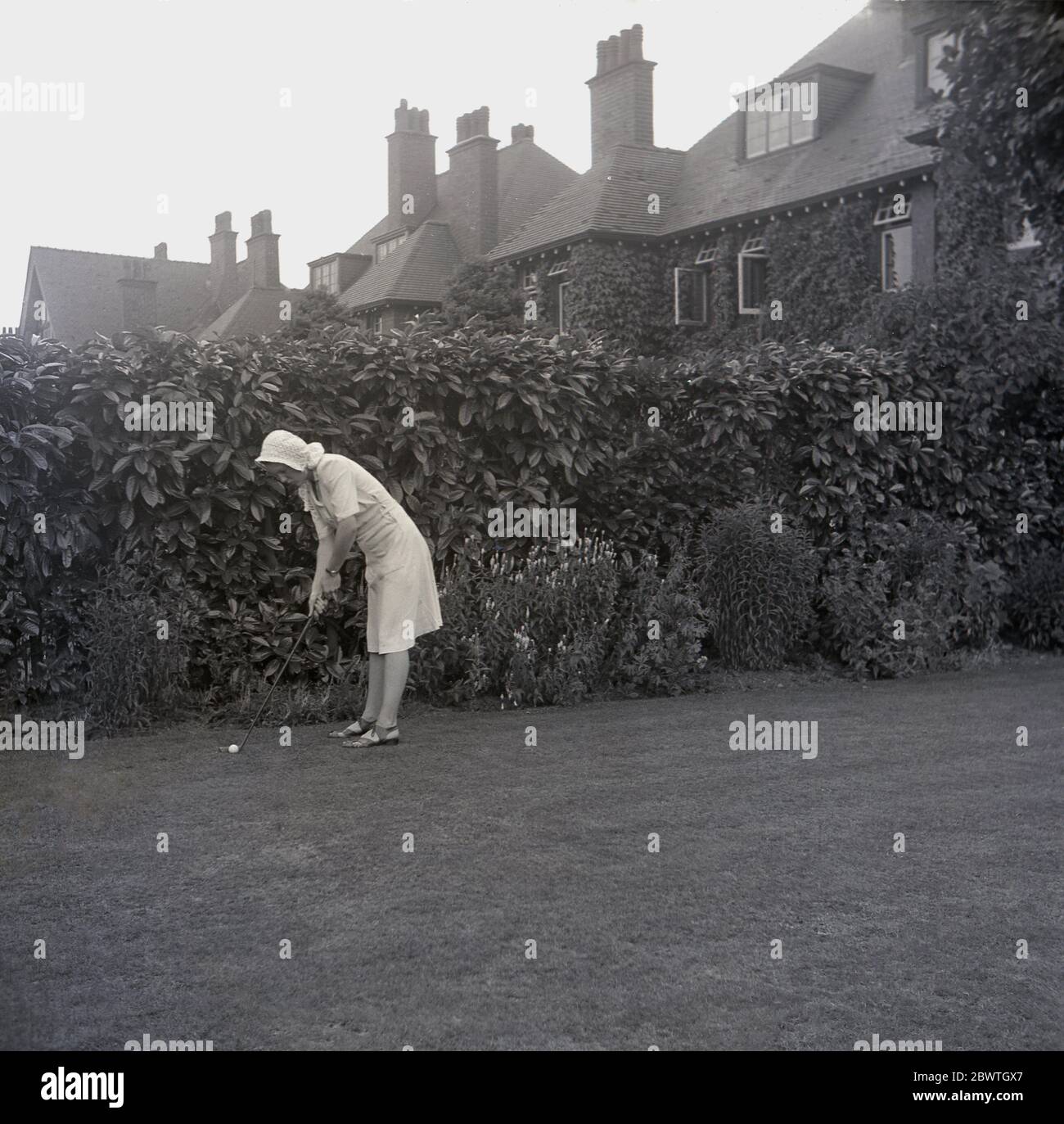 Anni 60, storico, in piedi accanto ad una siepe alta sul prato all'aperto in un giardino nel suburban Inghilterra, una giovane donna che indossa un cofano e dresss tenendo un putter tradizionale forma in piedi in piedi pronto a putt una palla da golf lungo l'erba. Foto Stock
