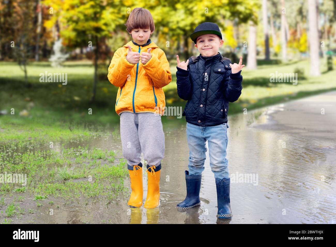 Due ragazzi allegri in stivali di gomma in piedi in puddle nel parco mostrando pollici verso l'alto gesto. Concetto di adattamento sociale dei bambini autistici Foto Stock