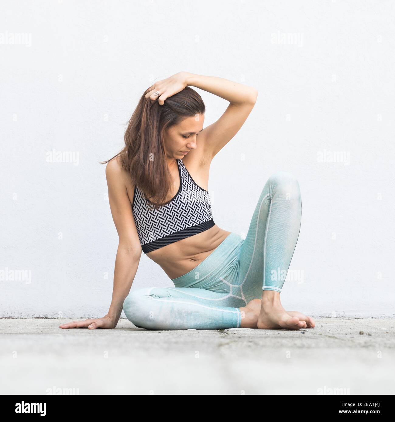 Montare sportivo ragazza attiva in sportswear seduto sul pavimento nella parte anteriore del muro grigio. Foto Stock