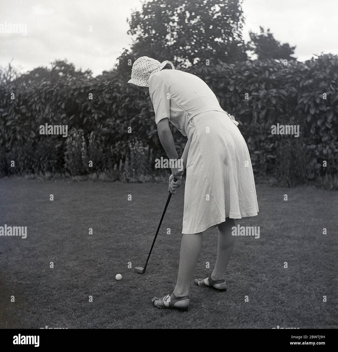 1960, storico, in piedi su un prato in un giardino di backgarden nella periferia dell'Inghilterra, una giovane donna che indossa un cofano e vestito con un putter tradizionale a forma di in piedi in piedi pronti a putt una palla da golf lungo l'erba. Foto Stock