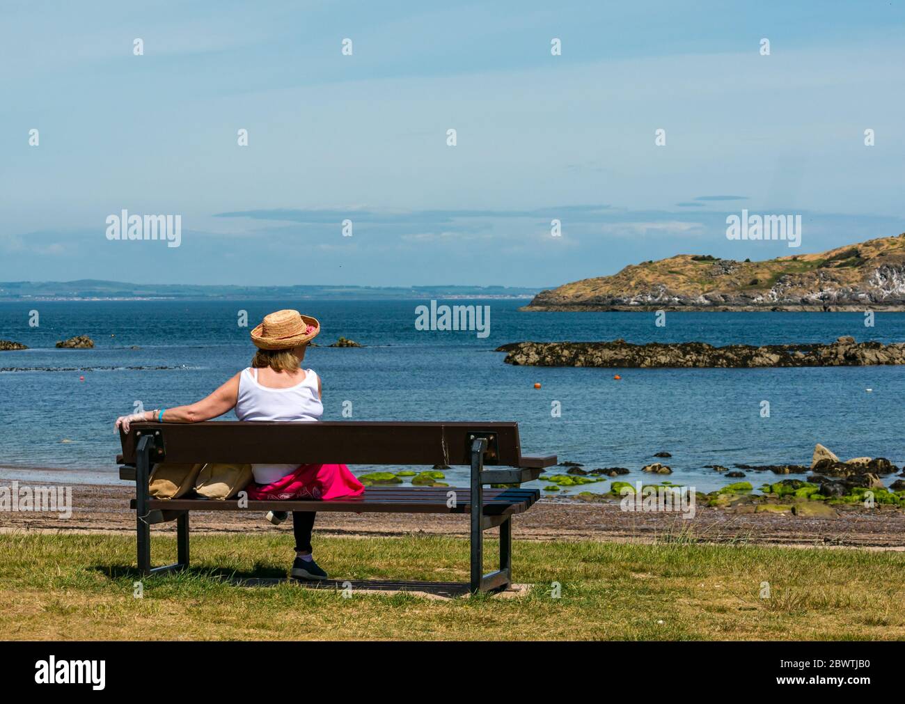 Donna che indossa cappello di paglia seduta su panca guardando la vista il giorno di sole, North Berwick, Scozia, Regno Unito Foto Stock