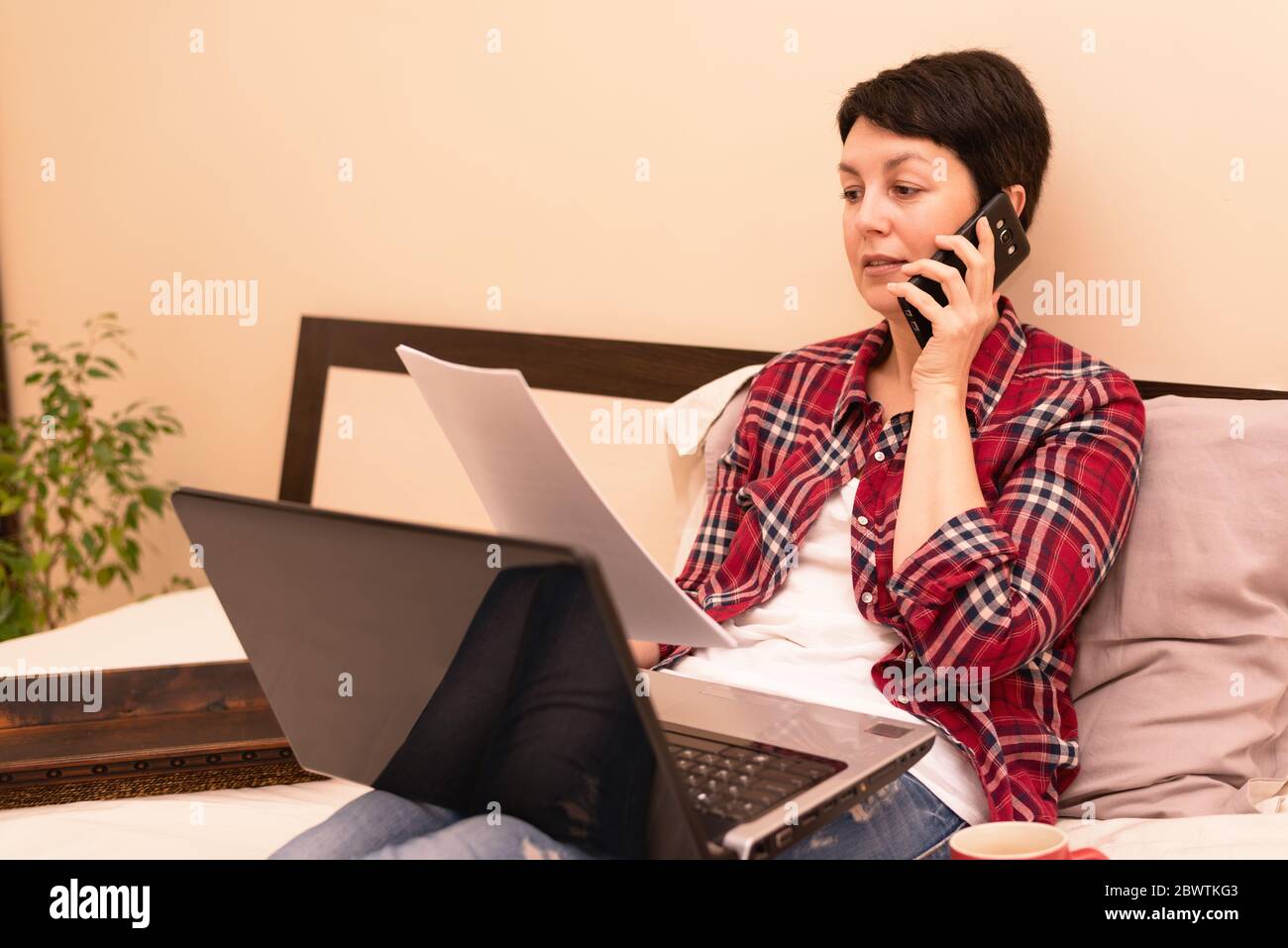 Donna caucasica che lavora a distanza da casa. Bruna dai capelli corti seduta sul letto usando il laptop e lo smartphone. Lavoro a distanza, concetto di lavoro freelance Foto Stock
