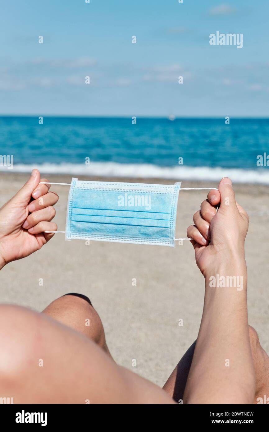 primo piano di un giovane caucasico con una maschera chirurgica blu nelle mani sulla spiaggia Foto Stock