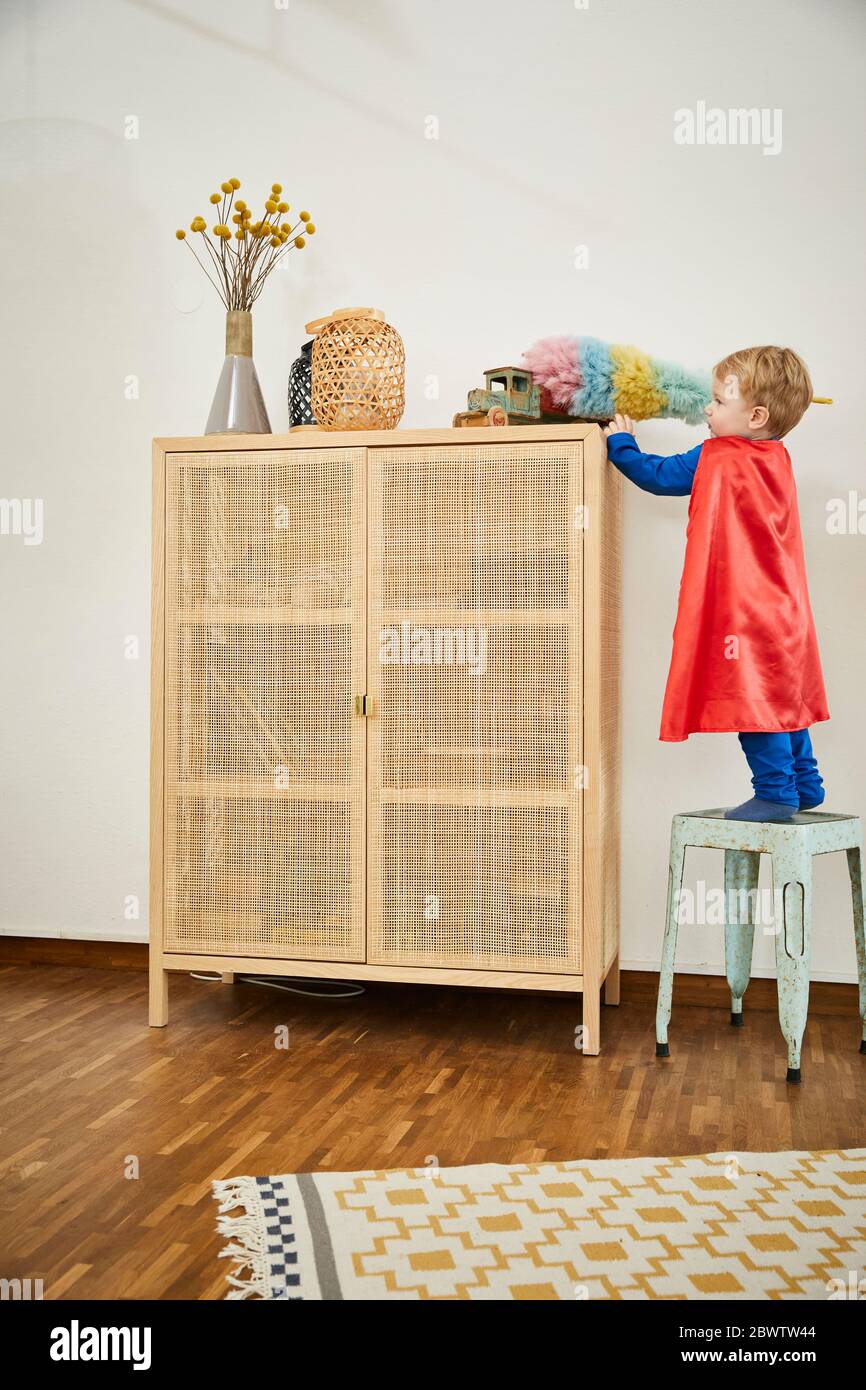 Ragazzo che indossa un costume superman e un giocattolo per la pulizia con un piumino su una sponda a casa Foto Stock