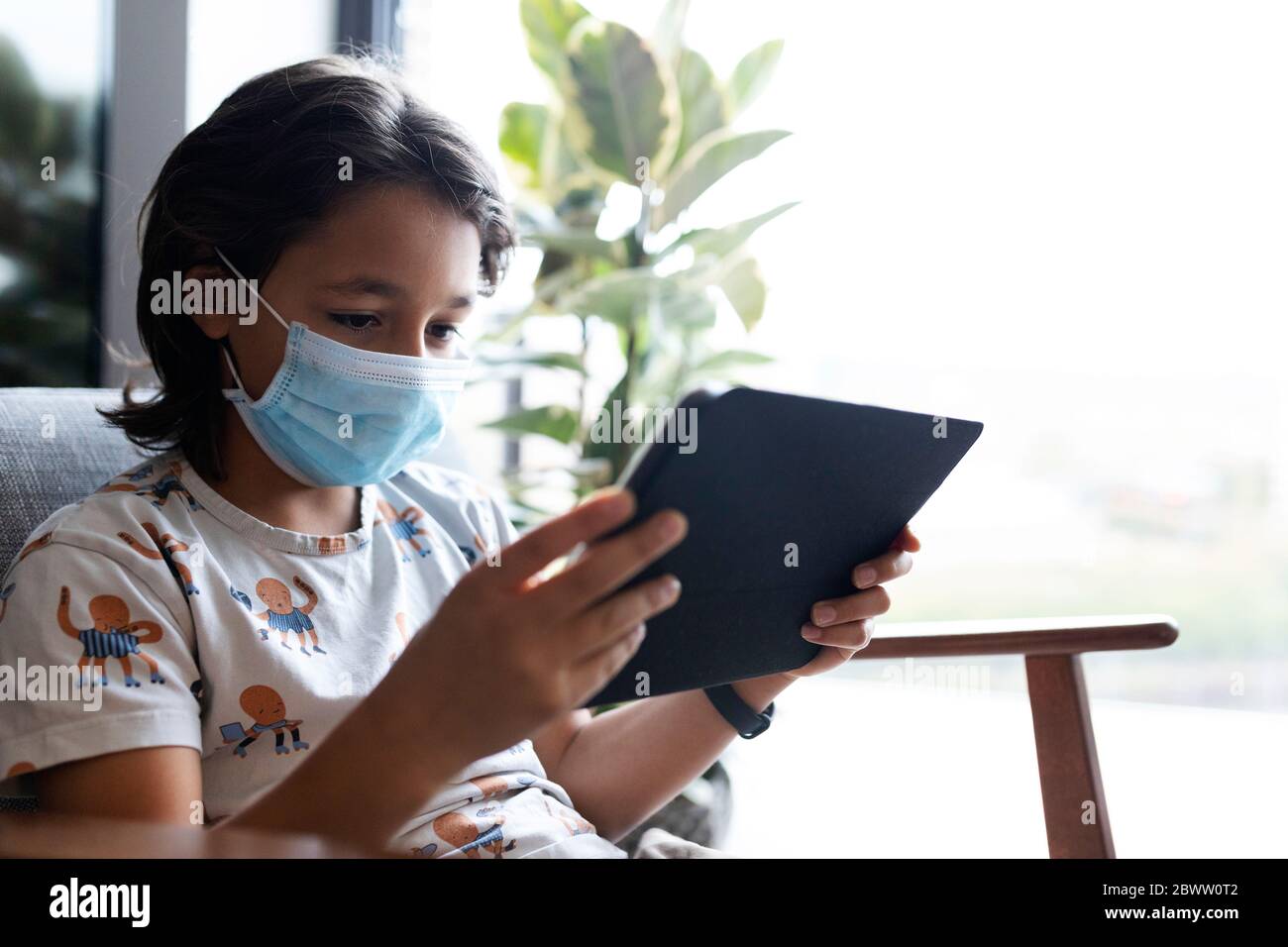 Ritratto di ragazzo con maschera chirurgica seduta su poltrona utilizzando tablet digitale Foto Stock