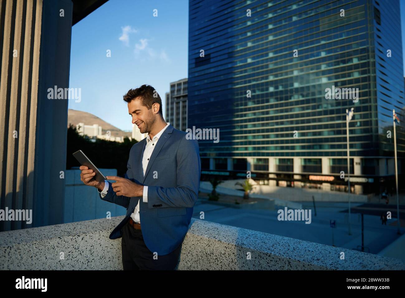 Uomo d'affari sorridente in piedi su una terrazza dell'ufficio usando un tablet Foto Stock