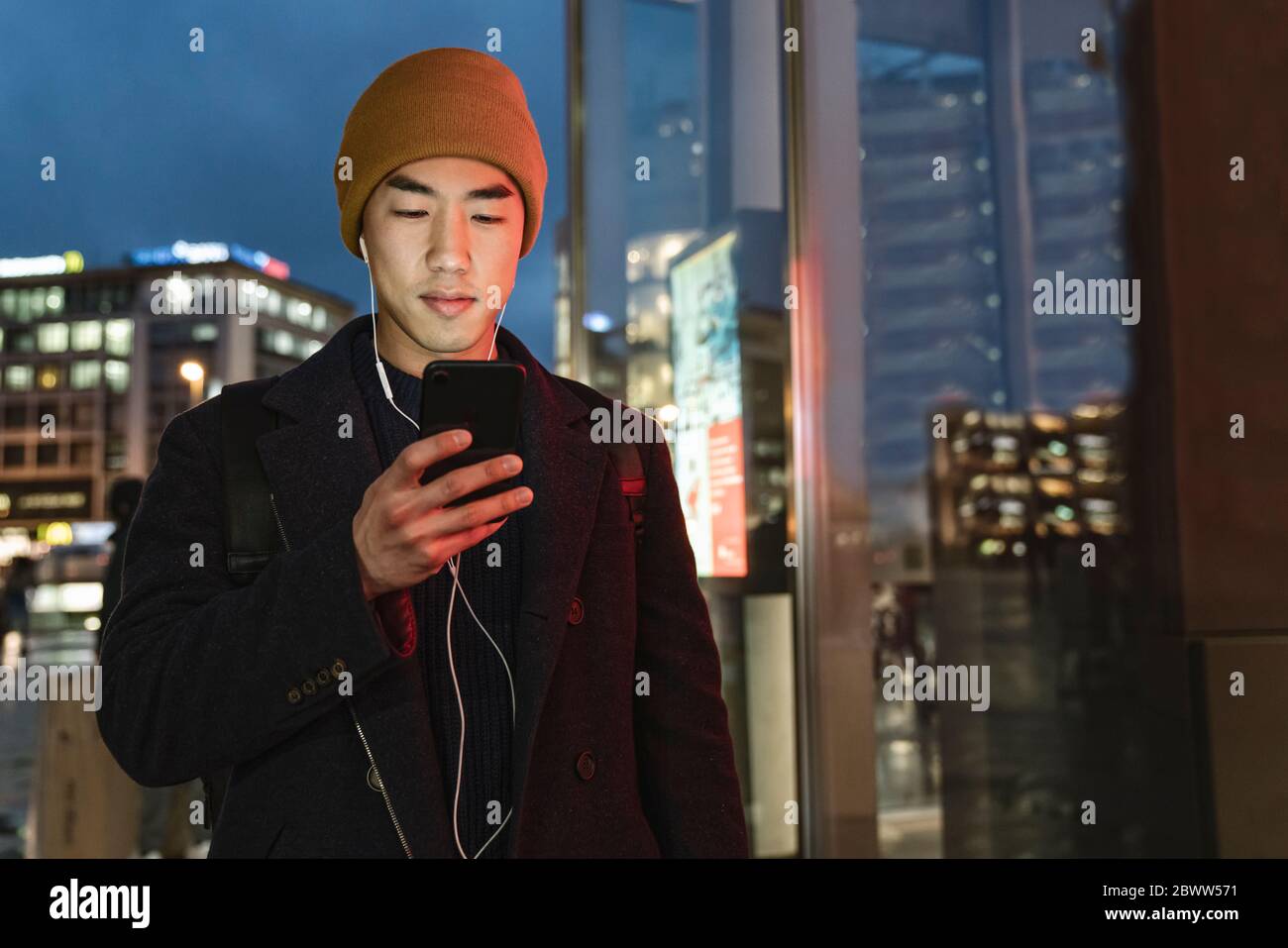 Uomo elegante con cappello giallo e auricolari che usano smartphone la sera in strada di notte Foto Stock