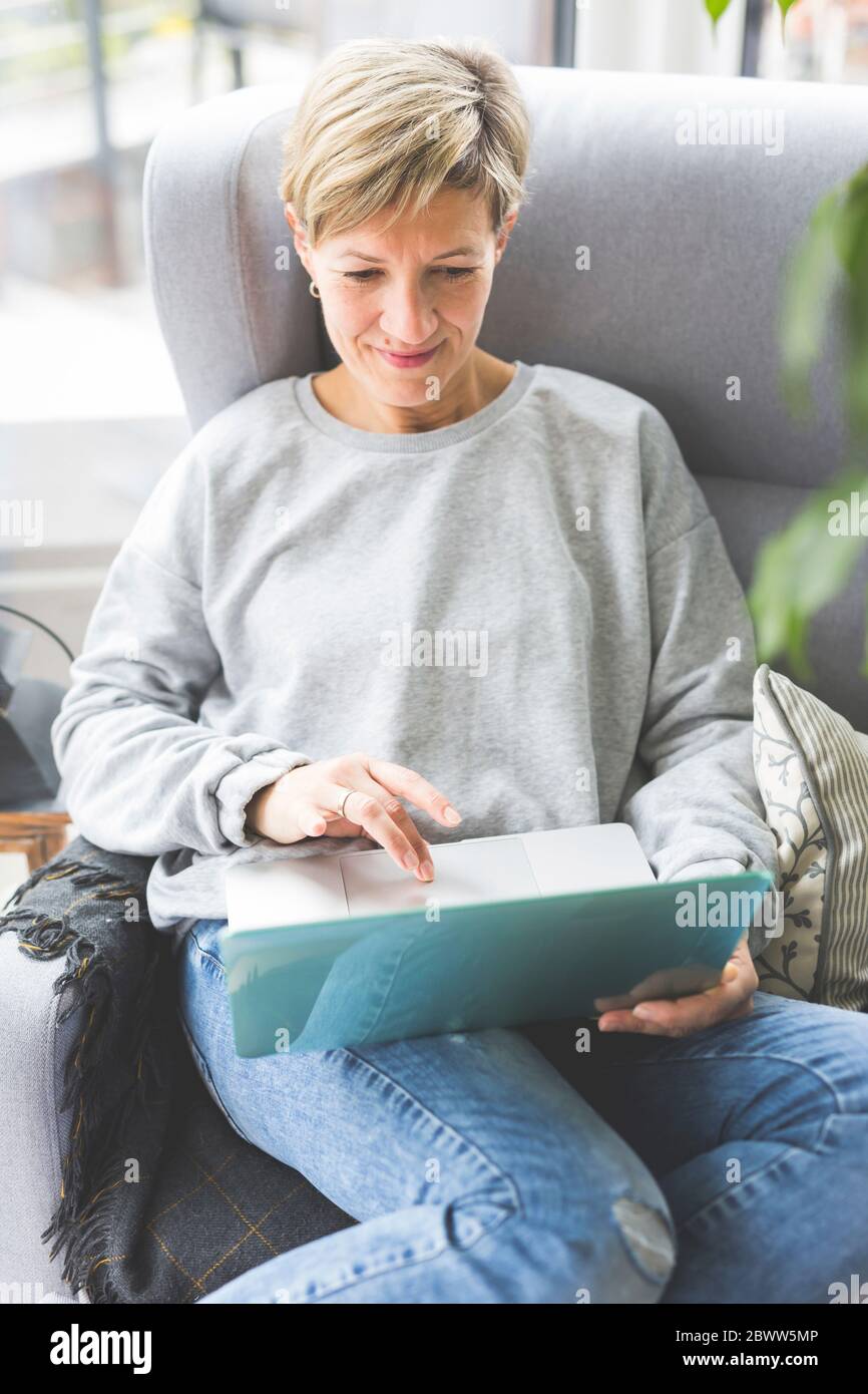 Donna matura che lavora da casa, con un computer portatile, seduta in poltrona Foto Stock