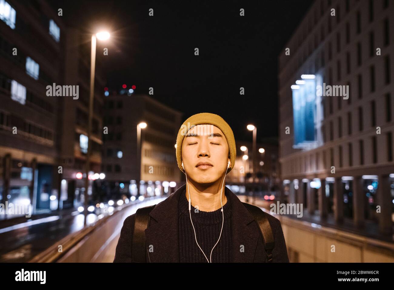 Ritratto di uomo elegante con cappello giallo e auricolari con occhi chiusi in città di notte Foto Stock