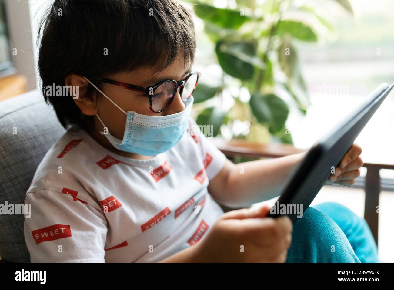 Ragazzo con maschera chirurgica seduta su poltrona utilizzando tablet digitale Foto Stock