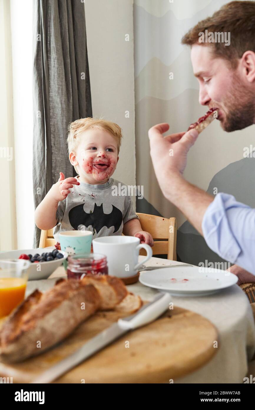 Ritratto di ragazzino sbavato al tavolo della colazione guardando suo padre mangiare pane con marmellata Foto Stock