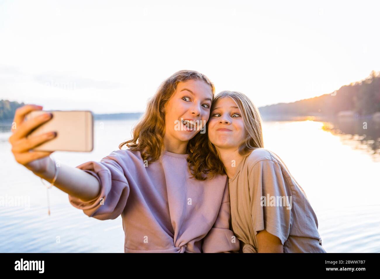 Amici felici che fanno i volti mentre si prendono selfie su smartphone contro lago Foto Stock