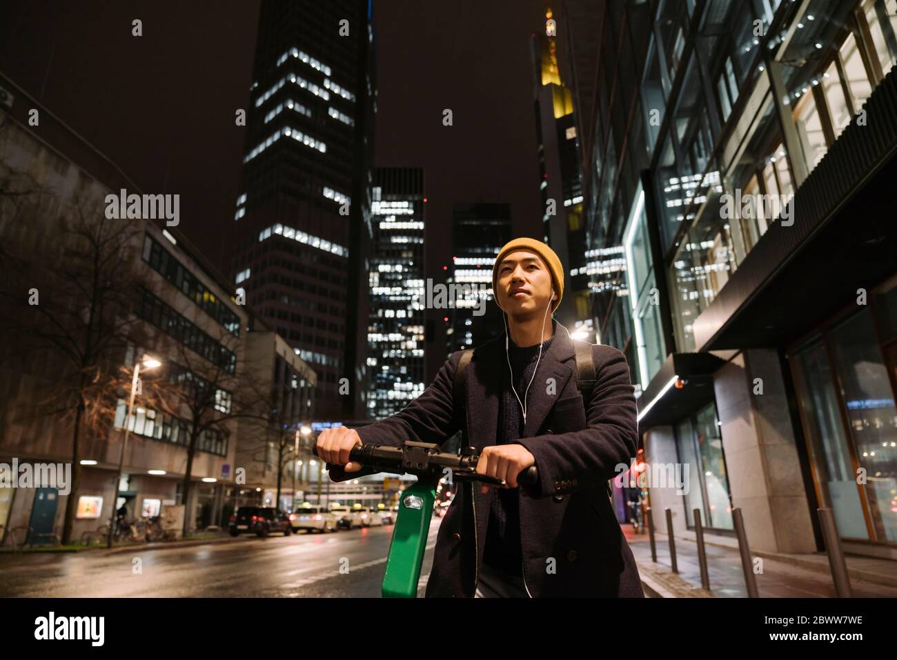 Uomo con cappello giallo e auricolari che usano scooter in città di notte Foto Stock