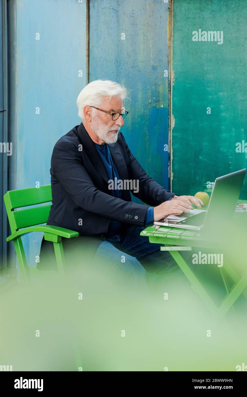 Uomo anziano sopportato seduto al tavolo da giardino usando il laptop Foto Stock