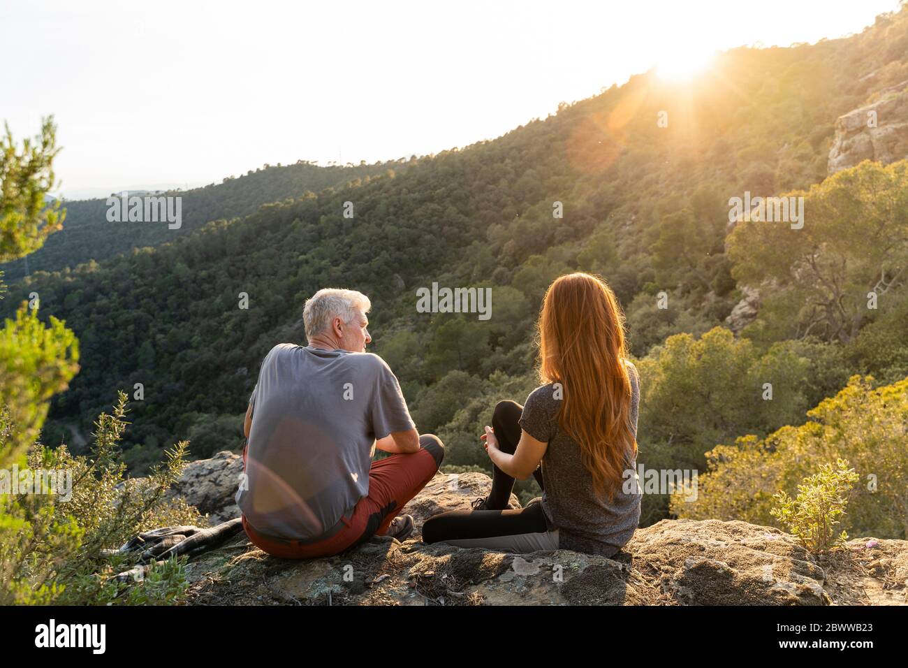 Vista posteriore di padre e figlia adulta seduta sulla roccia in montagna godendo il tramonto Foto Stock