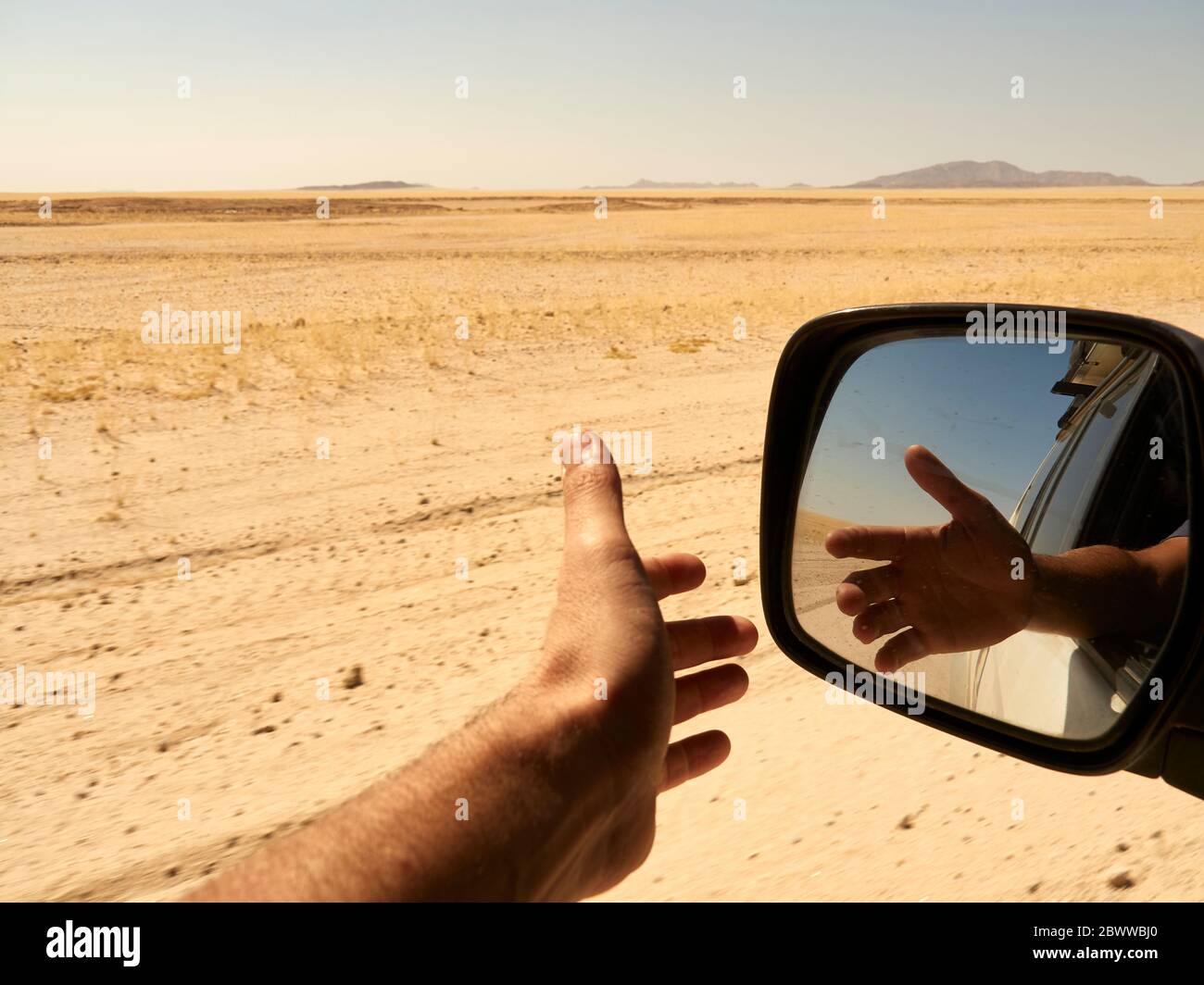 Riflesso nello specchio retrovisore della mano di un uomo fuori dal finestrino durante la guida nel deserto, Sossusvlei, Namibia. Foto Stock