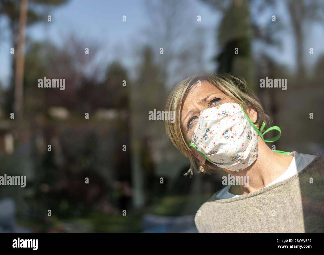 Donna con maschera facciale che guarda attraverso il vetro della finestra in giornata di sole durante la quarantena Foto Stock