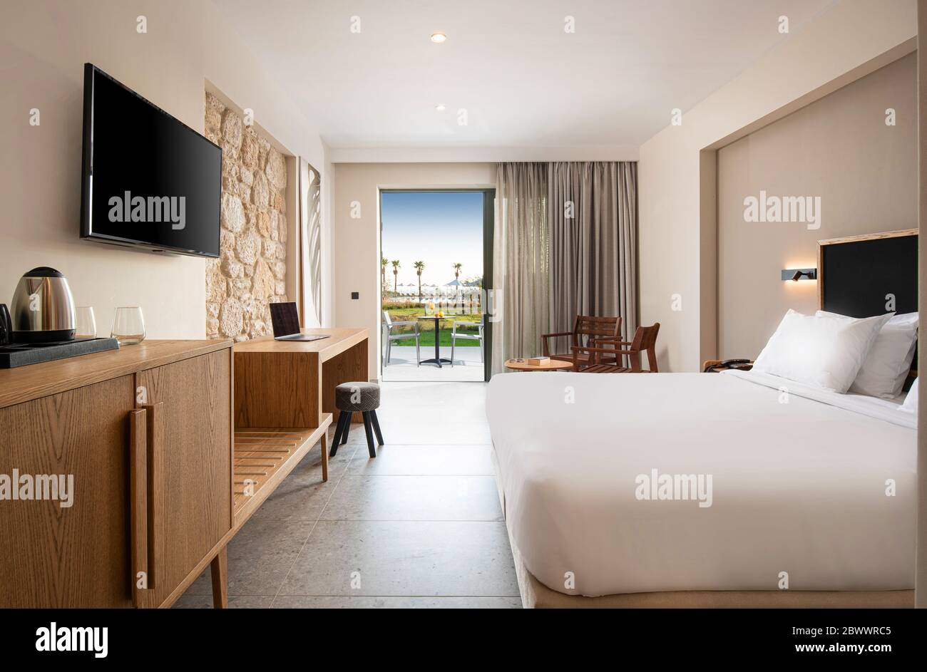 Interni luminosi e vuoti della camera da letto contemporanea dell'hotel con pareti in pietra beige, mobili in legno e terrazza all'aperto Foto Stock