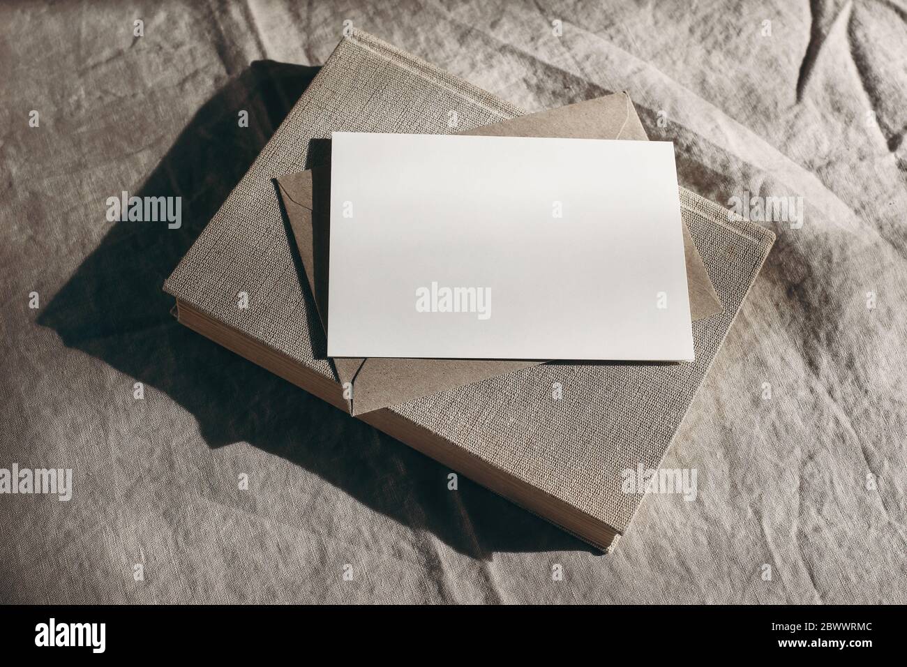 Moody Stationery ancora scena di vita. Libro vecchio, foglio bianco di carta, mockup di biglietti d'auguri e busta alla luce del sole. Sfondo tovaglia di lino beige Foto Stock