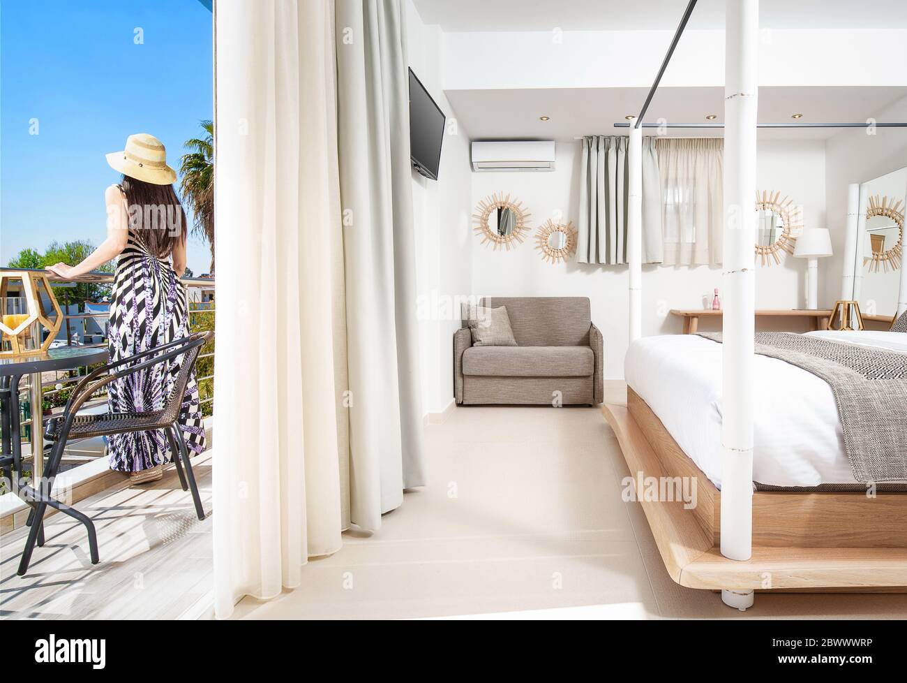 Giovane donna in stile romantico vestito in piedi sul balcone della moderna camera da letto dell'hotel resort. Interno luminoso a spazio aperto con arredi in legno bianco, grigio Foto Stock