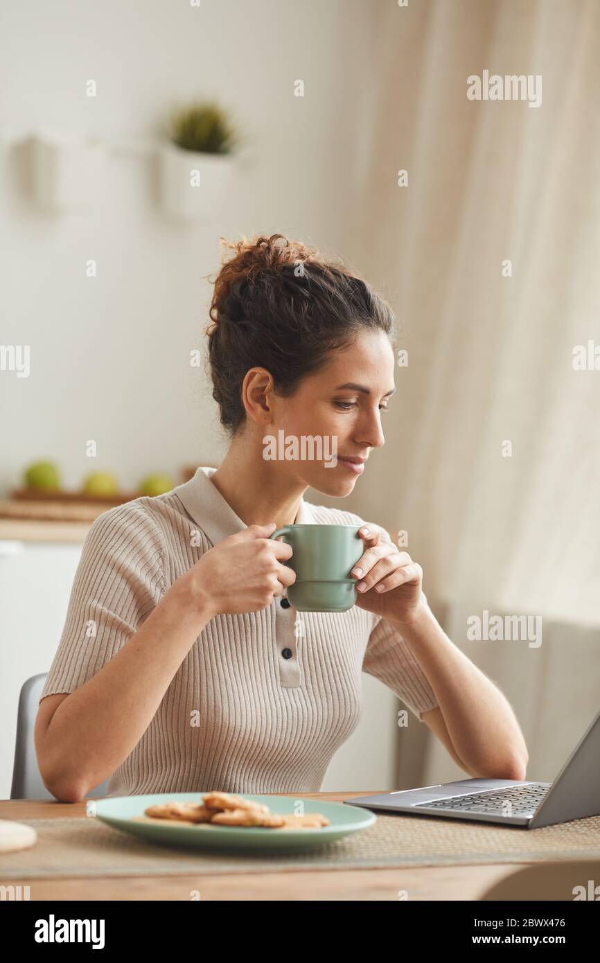 Giovane donna seduta al tavolo e bevendo caffè mentre si usa il computer portatile a casa Foto Stock