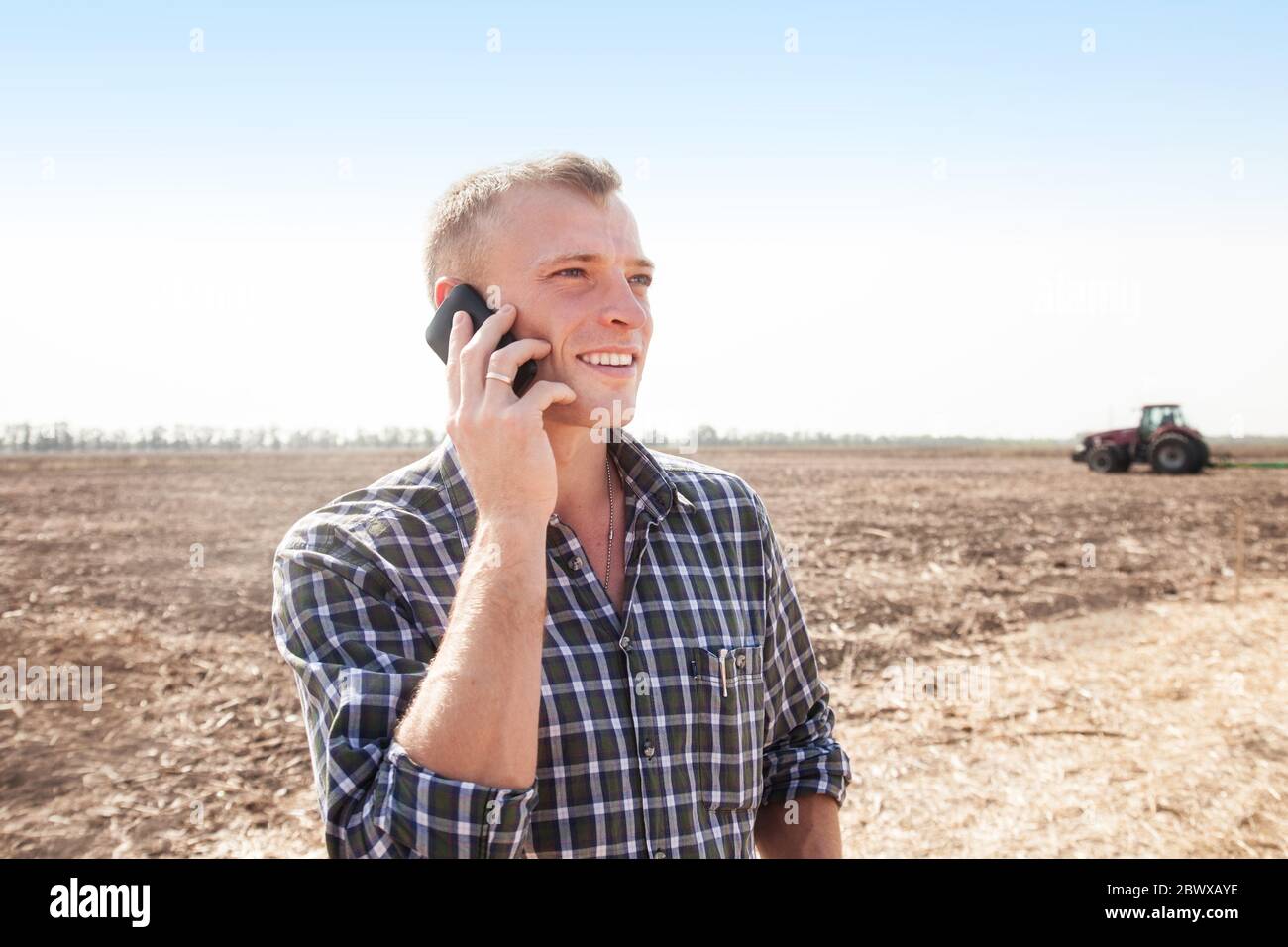 Giovane uomo attraente con telefono vicino a un trattore. Concetto di agricoltura e lavori sul campo. Foto Stock