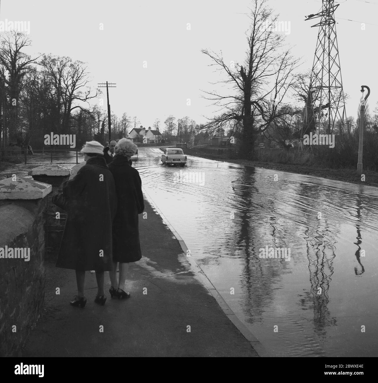 1960, storico, un'auto dell'epoca che scende su una strada allagata come due Signore in cappotti si trovano su un marciapiede preoccupato che stanno per ottenere schizzato, Inghilterra, Regno Unito. Foto Stock
