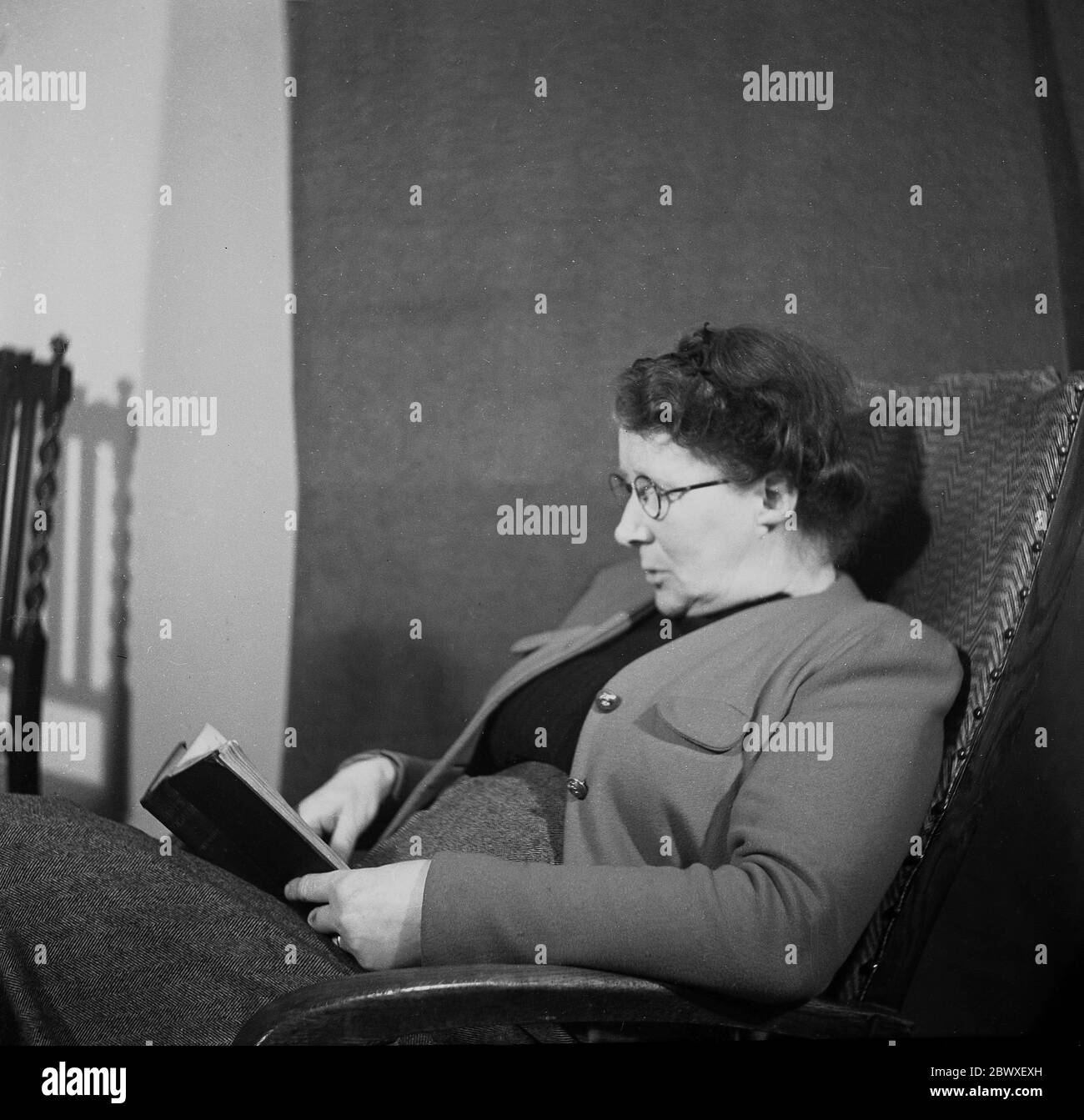 Anni '50, storica, una signora inglese seduta in una sedia reclinabile in legno incorniciato che legge un libro, Inghilterra, Regno Unito. Foto Stock