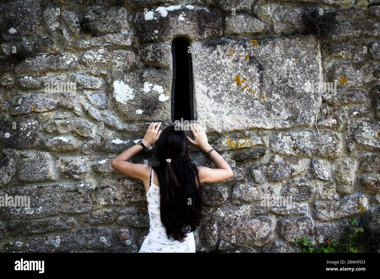 Donna curiosa che cerca di salire su un muro di pietra per guardare attraverso una finestra Foto Stock