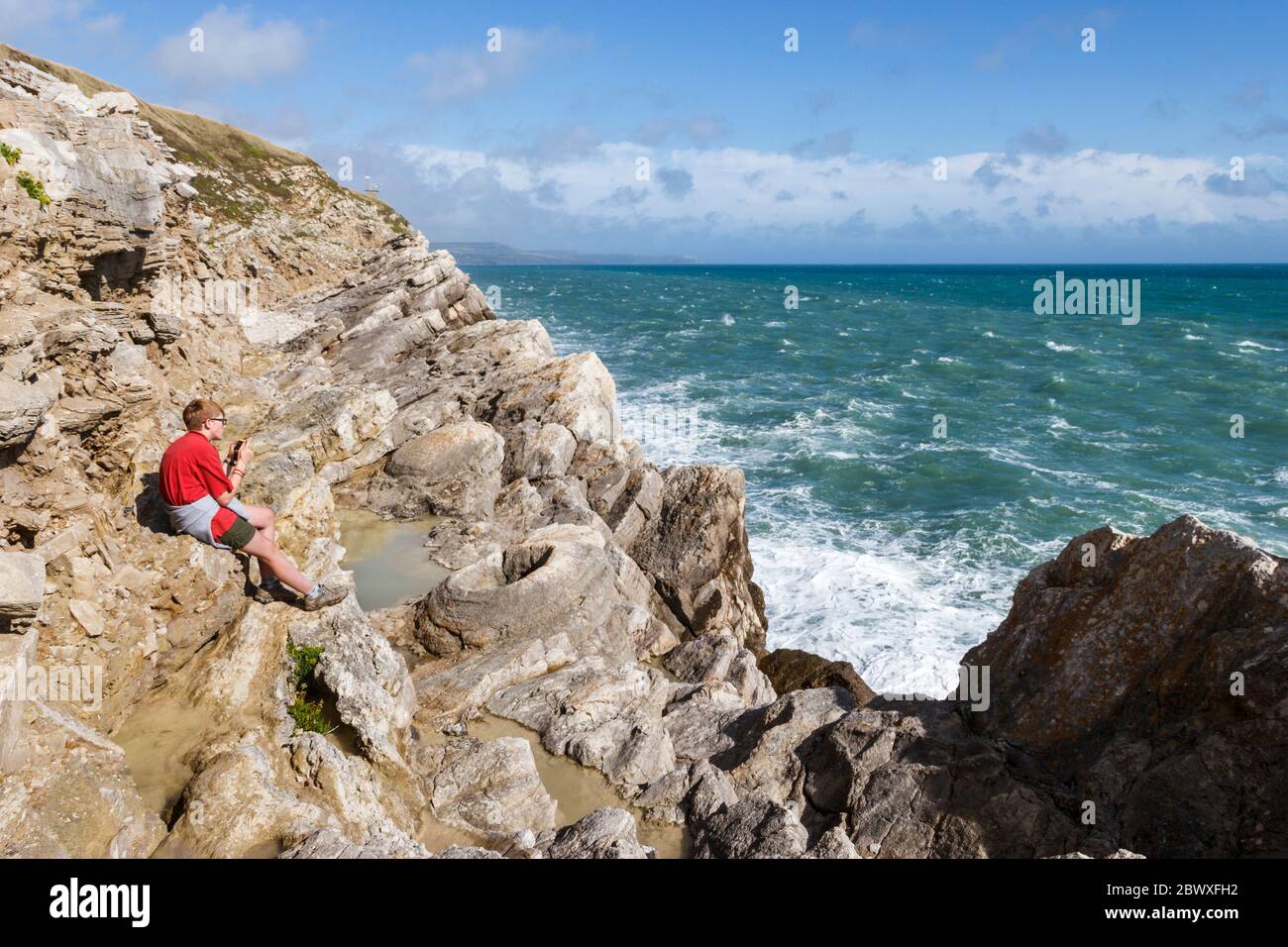 Una ragazza di quindici anni in una camicia rossa seduta su rocce e scattando foto nella foresta fossile sulla costa vicino a Lulworth, Dorset, Regno Unito Foto Stock