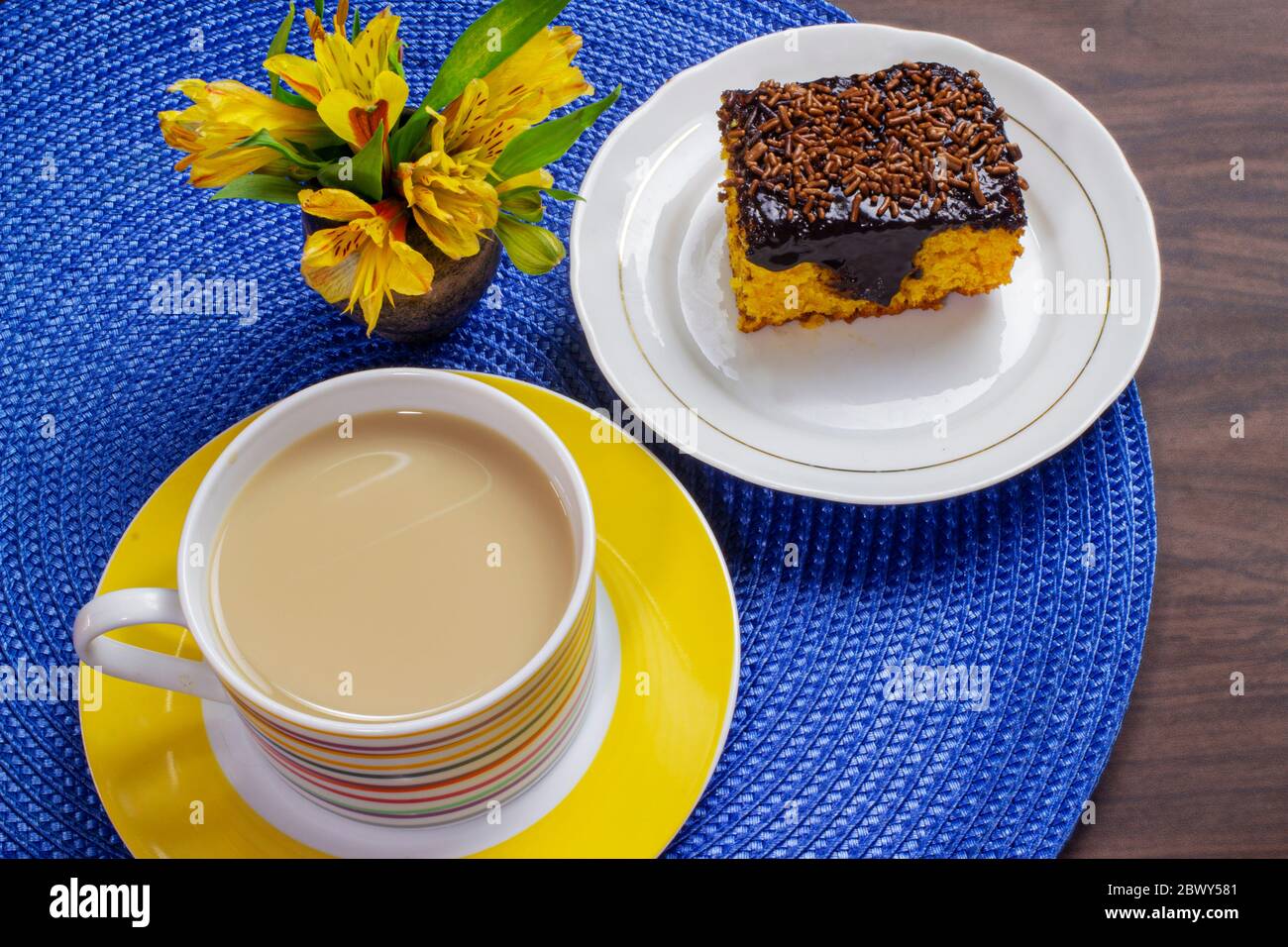 Torta di carote con glassa di cioccolato e tazza di latte con caffè su tavolo di legno decorato con un fiore giallo. Foto Stock