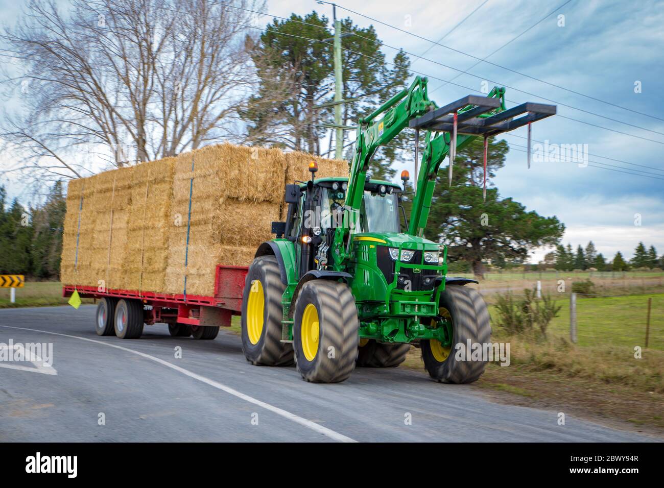 Arundel, Canterbury, Nuova Zelanda, 3 giugno 2020: Un trattore agricolo sposta un carico di rimorchio di balle di paglia lungo una strada di campagna in inverno per l'alimentazione di stock Foto Stock