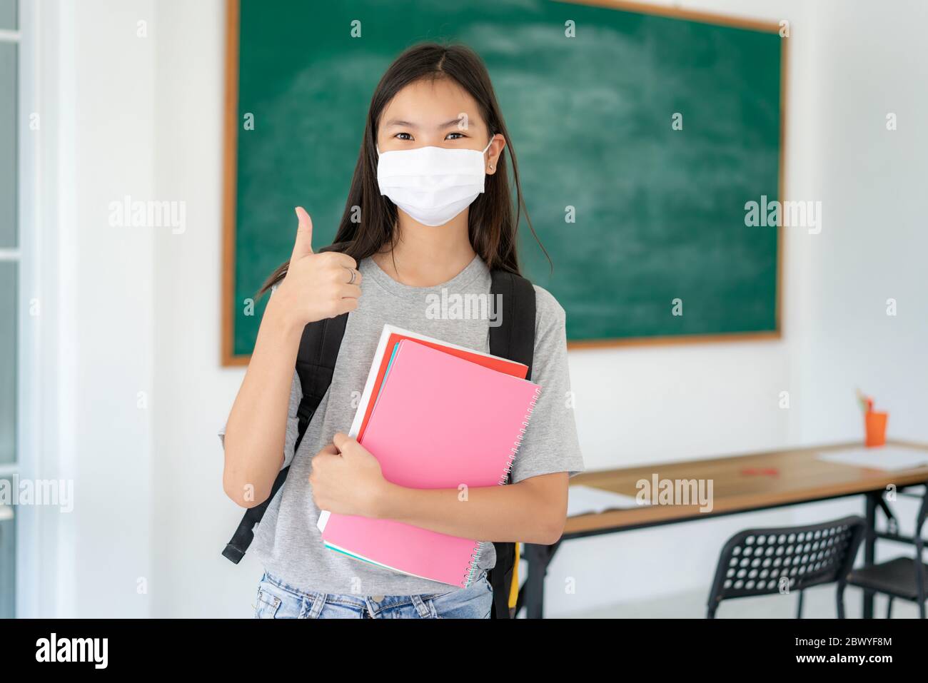 Gli studenti asiatici primari ragazza pollice su e indossare maschere per prevenire lo scoppio di Covid 19 in classe, mentre di ritorno a scuola riaprire la loro scuola, New Foto Stock