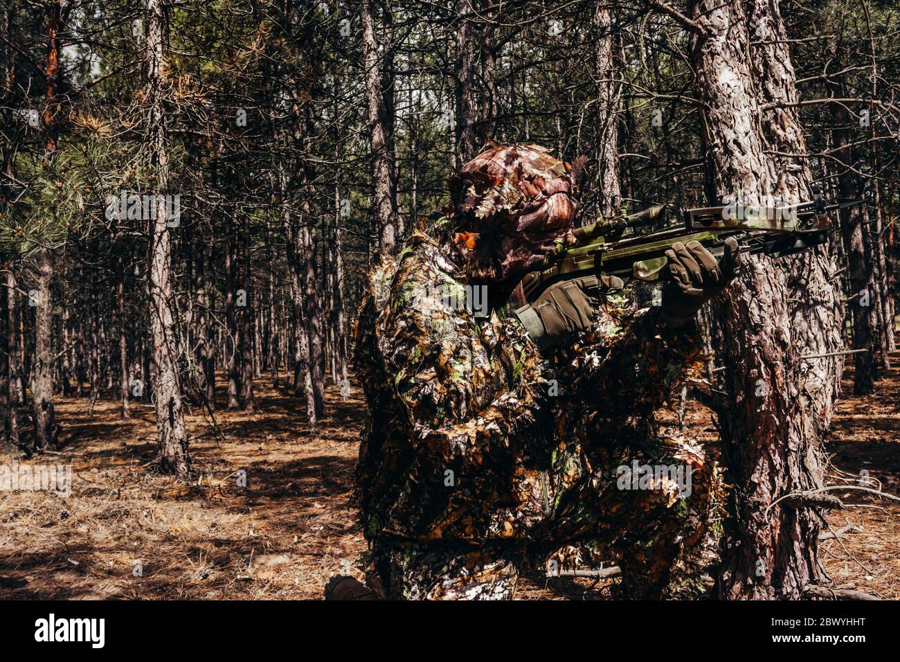 Foto di un cacciatore di cecchini mimetizzato con balestra seduta in boschi. Foto Stock