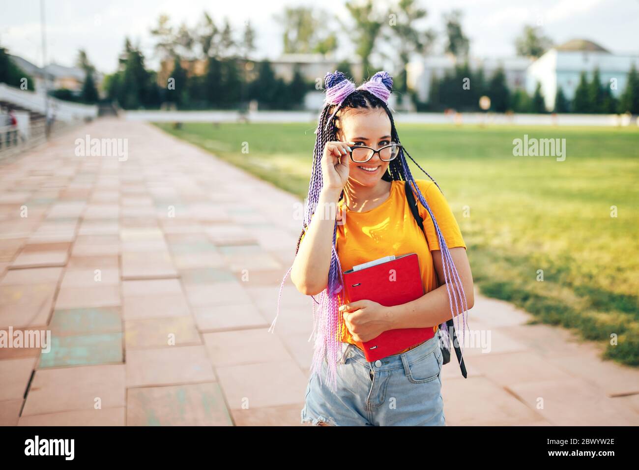 Giovane bella ragazza che tiene una cartella rossa in mano in una T-shirt gialla che cammina all'aperto in uno stadio sportivo ragazza studentesca di nuovo a scuola Foto Stock