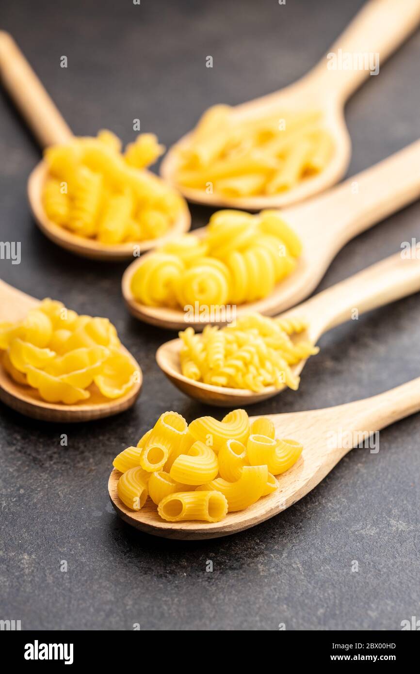 Vari tipi di pasta italiana cruda in cucchiai di legno su tavola nera. Foto Stock