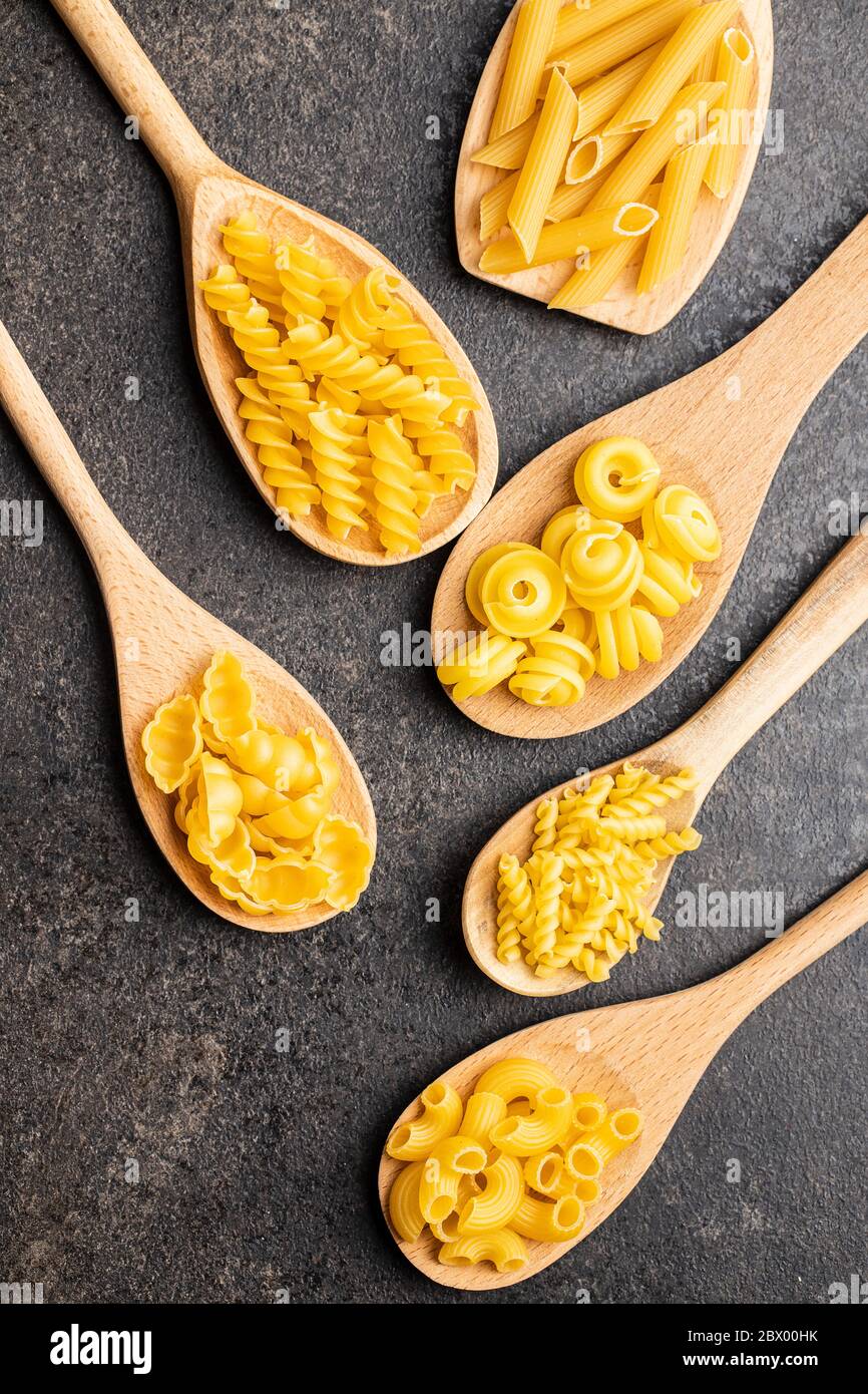 Vari tipi di pasta italiana cruda in cucchiai di legno su tavola nera. Vista dall'alto. Foto Stock