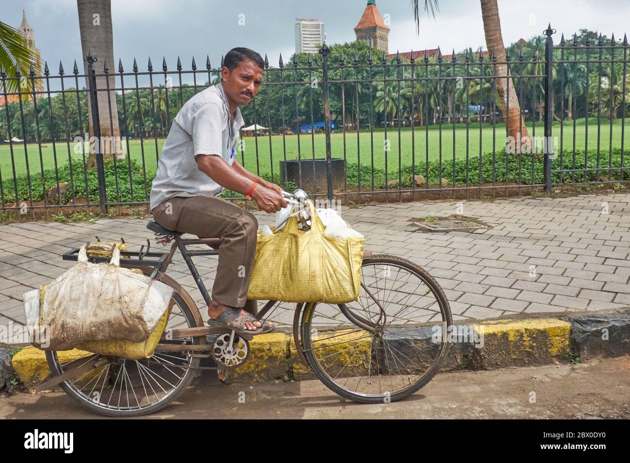 Un uomo a Mumbai, India, passa il verde di Oval Maidan, borse appese alla sua bicicletta, la Bombay Hight Court & Bombay Borsa nel b/g. Foto Stock