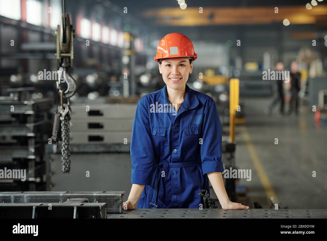 Ritratto di sorridente giovane specialista asiatico della lavorazione dei metalli in tute blu in piedi alla scrivania del negozio industriale Foto Stock