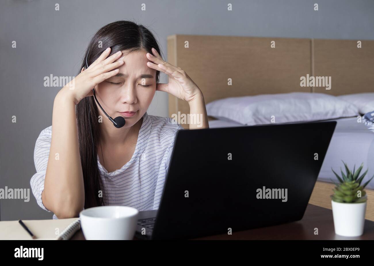 Donna asiatica che lavora a casa con cuffie che fanno videoconferenza con i colleghi tramite computer portatile con stress emotivo durante la trasmissione COVID-19 Foto Stock