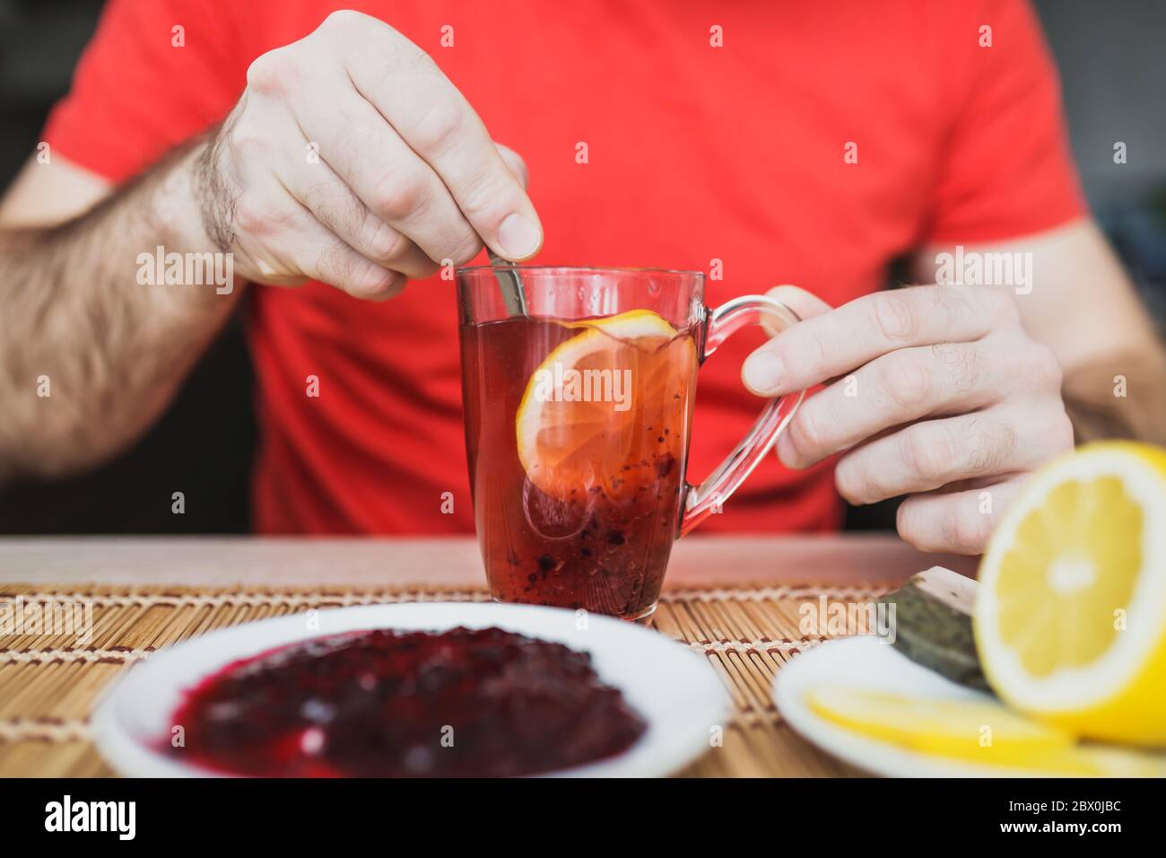 Primo piano di una mano dell'uomo che mescola limone e marmellata in una tazza di tè - una bevanda calda vitaminica utile per i raffreddori Foto Stock