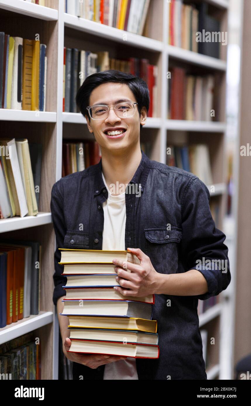 Giovane uomo cinese in occhiali in piedi in biblioteca con libri Foto Stock