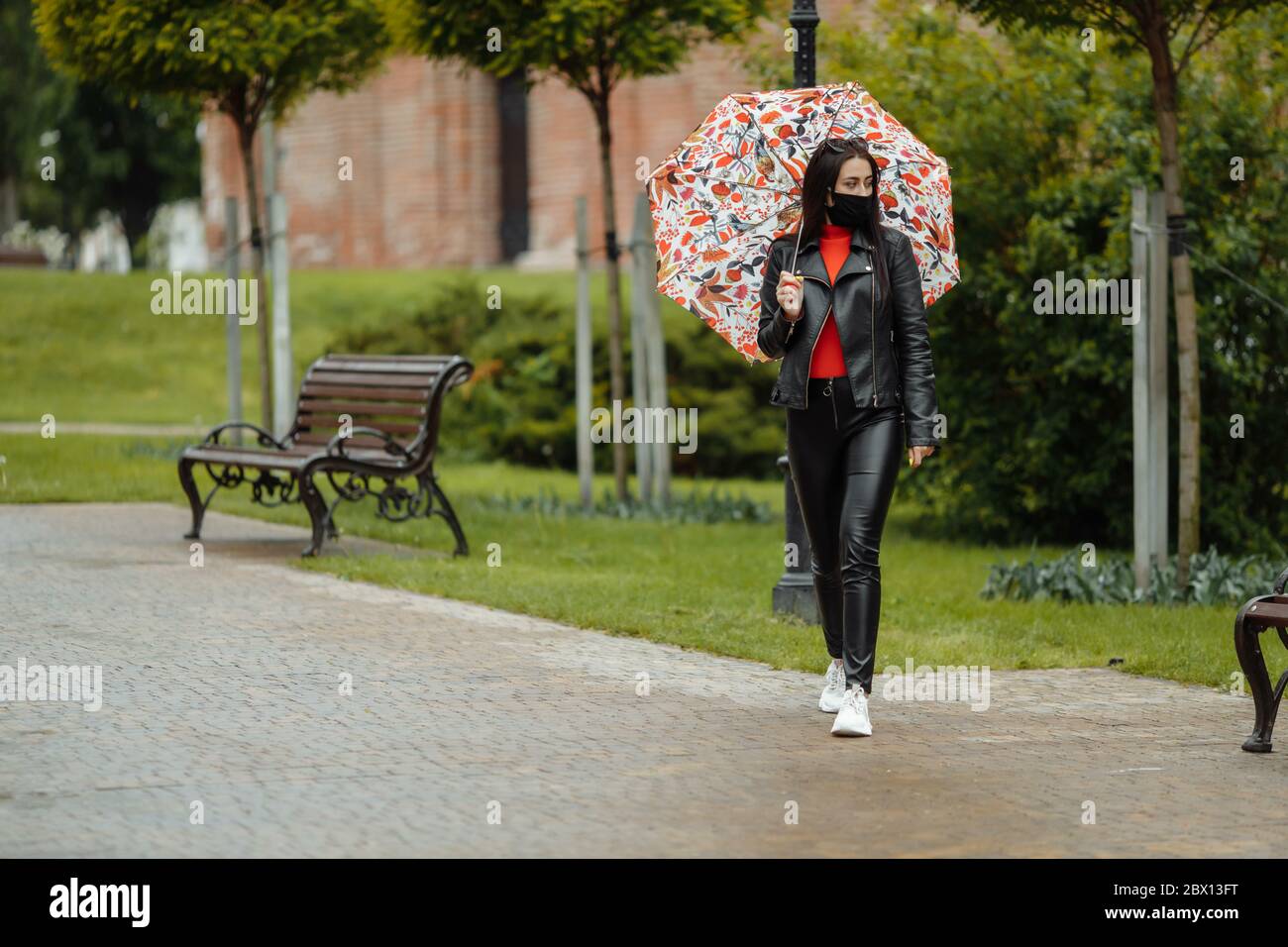 Una ragazza mascherata sta camminando lungo la strada. Una ragazza in una maschera protettiva cammina nel parco con un ombrello sotto la pioggia. Infezione da coronavirus COVID-19 Foto Stock