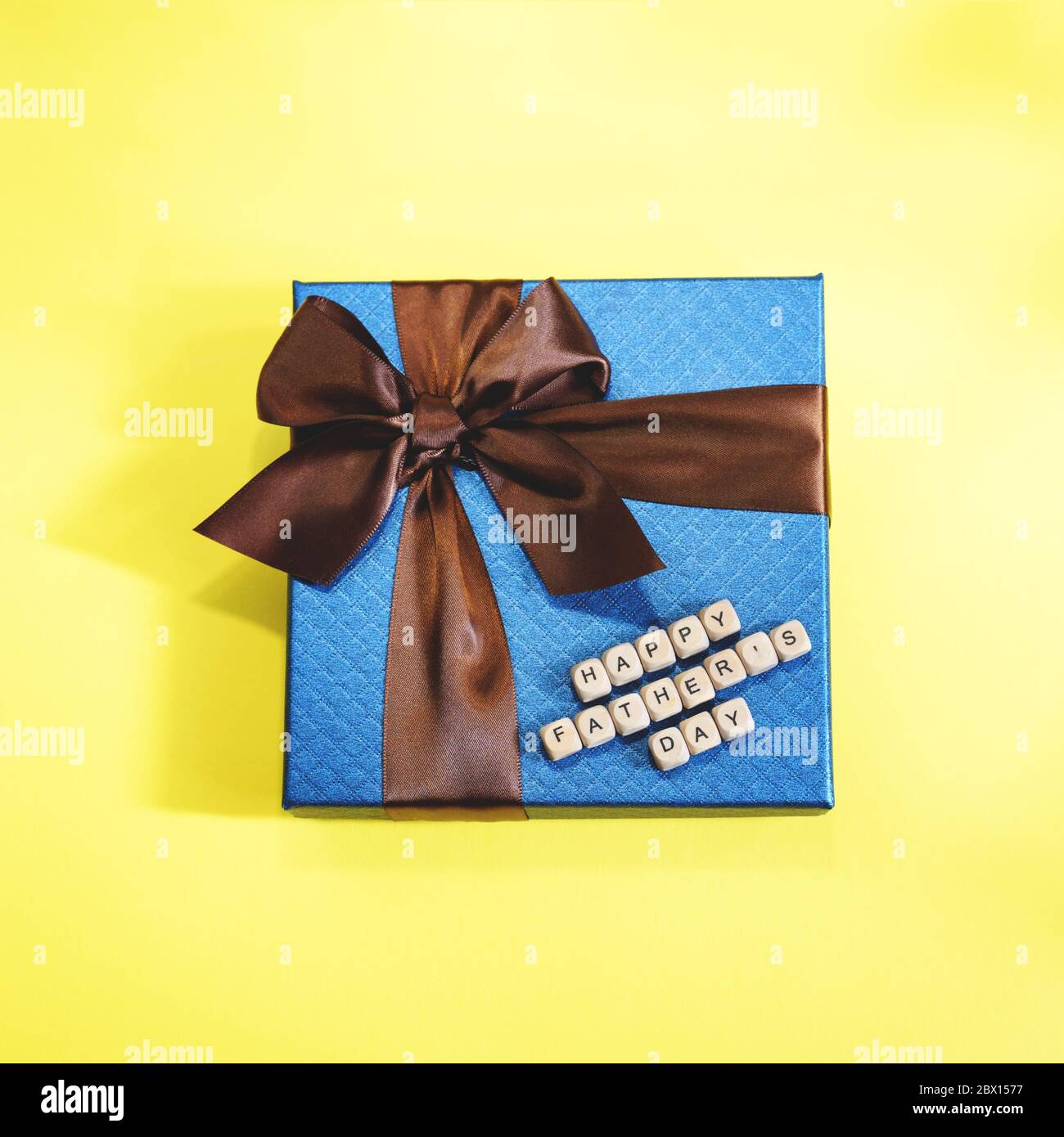 Un regalo blu con striscia marrone su sfondo giallo. Un segno di buon giorno del Padre da blocchi di legno. Concetto di festa del Padre. Foto Stock