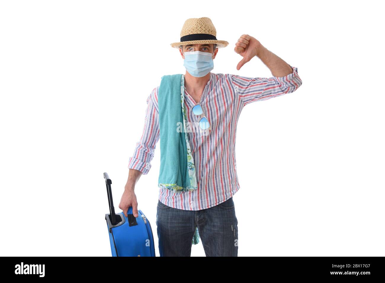 Uomo preparato per la vacanza con il pollice in giù protetto con maschera facciale, cappello di paglia, asciugamano, bicchieri e valigia in mano isolato con sfondo bianco Foto Stock