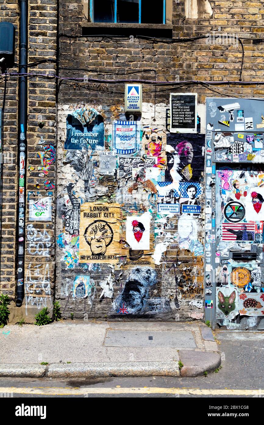 Muro in mattoni coperto di graffiti, etichette e poster in Fashion Street, Spitalfields, Londra, Regno Unito Foto Stock